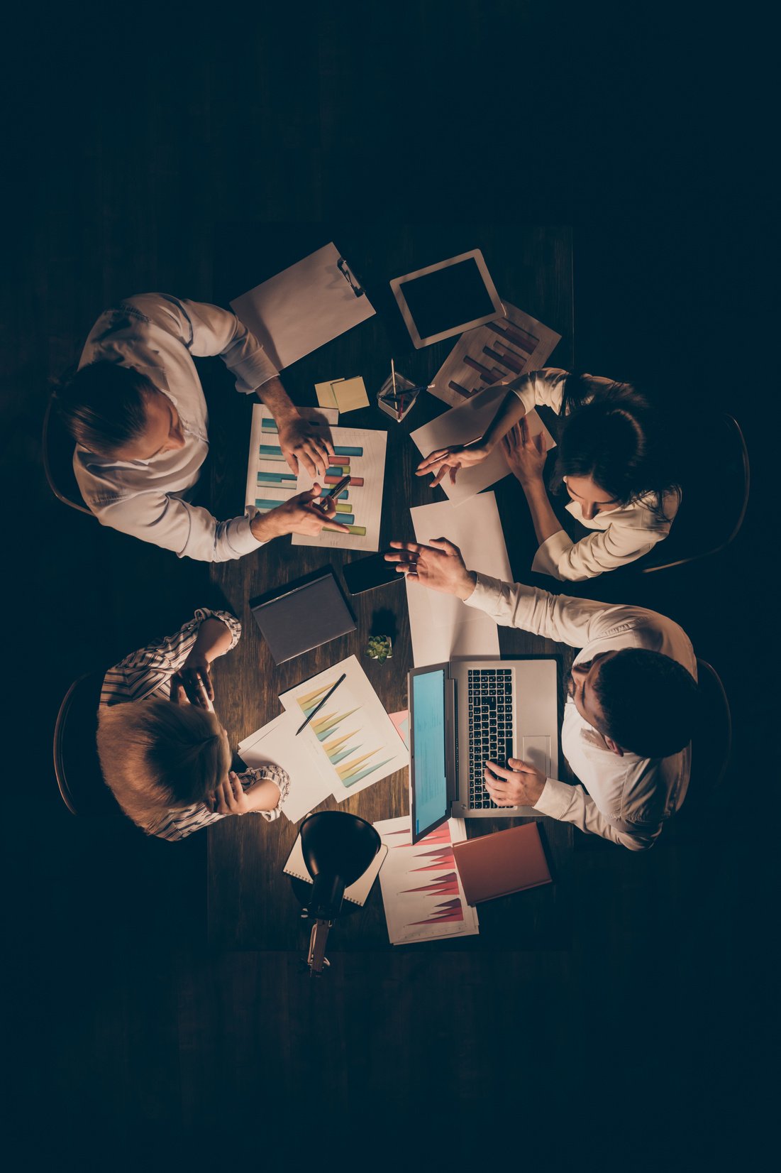 High angle above view vertical photo of four business partners sitting opposite working late night speak about new startup details hold schemes formalwear lamp light indoors